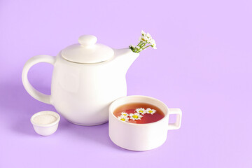 Teapot with cup of natural chamomile tea, sugar and flowers on lilac background