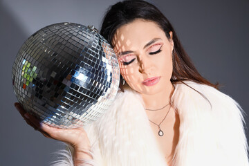 Fashionable beautiful young woman in fur coat with disco ball on grey background