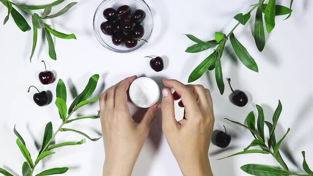woman apply cream to her hands, Branch with fresh green Ruscus leaves and cherry fruits on gray background