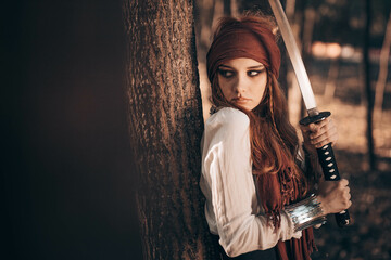 Young female in pirate costume with a sword leaning on the tree