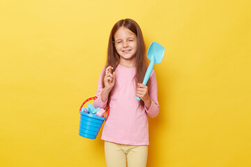 Hopeful little girl wearing casual clothing holding sandbox toys and bucket isolated over yellow background crossing fingers wants to go to  play outdoor in sand with puddle.