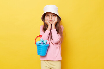 Funny little girl wearing casual clothing and panama holding sandbox toys and bucket looking at...