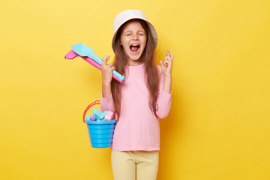 Cheerful joyful happy little girl wearing casual clothing and panama holding sandbox toys and bucket isolated over yellow background crossing fingers making wish.