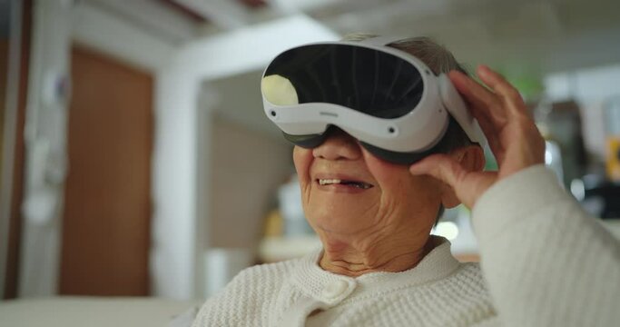 Happy senior asian woman sitting on sofa and playing VR. She looking social content in VR world and laughing in living room.