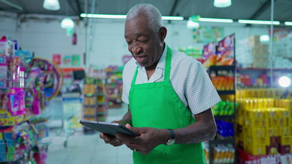 African American senior man employee of grocery store speaking on video with tablet device standing inside supermarket, communicating with staff. checking inventory