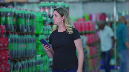 Female consumer walking through supermarket soda drinks aisle holding cellphone browsing products and looking at grocery list. Woman shopping depicting consumerism lifestyle habits