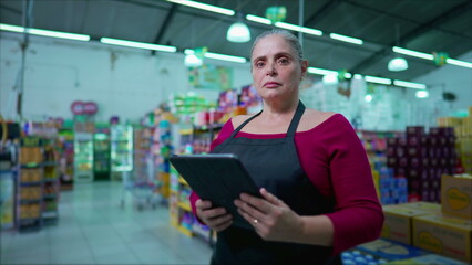 Serious female middle-age employee of supermarket standing inside business holding tablet device,...
