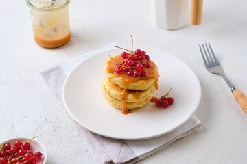 Breakfast, portioned cottage cheese pancakes with salted caramel and red currants on a white plate on a light concrete background.