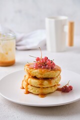 Breakfast, portioned cottage cheese pancakes with salted caramel and red currants on a white plate on a light concrete background.