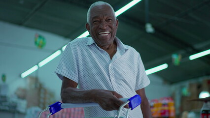 One happy Brazilian senior shopper smiling at camera with shopping cart inside supermarket shed....