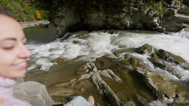 woman tourist sitting on rocks next to mountain river with waterfalls. female hiker taking photo and video using phone camera during autumn mountain hike. outdoor active leisure, enjoying nature