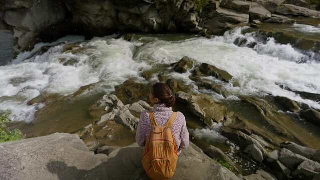 woman tourist sitting on rocks next to mountain river with waterfalls. female hiker taking photo and video using phone camera during autumn mountain hike. outdoor active leisure, enjoying nature