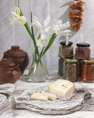 A piece of cheese cut on a wooden board on a light table. There are irises in a transparent vase. Spring