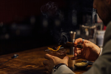 Man smoking a cigar and having an old-fashioned cocktail served on a wooden bar table