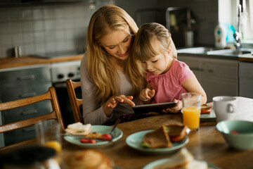 Mother and daughter using a digital tablet while having breakfast in the kitchen in the morning