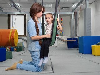 Child with physiotherapist on swing during sensory integration session. Little girl in...