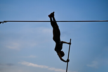 A female pole vaulter (silhouette) jumping with a beautiful blue sky in the background. Track and field athlete. Young woman pole-vaulting. Pole vault competition - obrazy, fototapety, plakaty