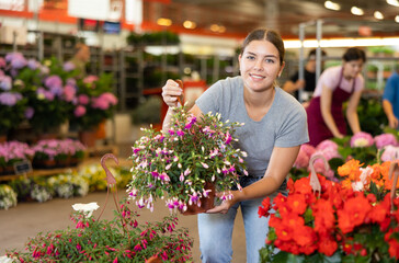 Fototapeta na wymiar Glad young woman purchasing fuchsia in garden pot in point of sale of plants outdoors