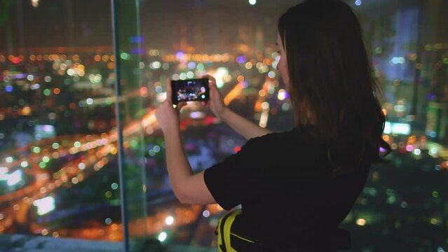 Tourist woman shoot night cityscape aerial view panorama with mobile phone standing on observation deck. Girl takes pictures of city lights on smartphone. Top floor of business center tower viewpoint