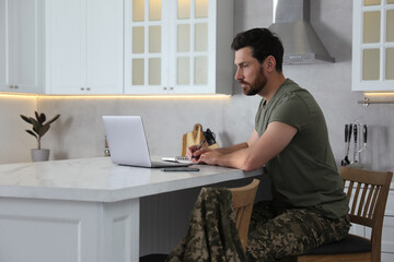 Soldier taking notes while working with laptop at white marble table in kitchen. Military service
