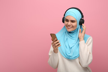 Muslim woman in hijab and headphones listening to music with smartphone on pink background, space for text