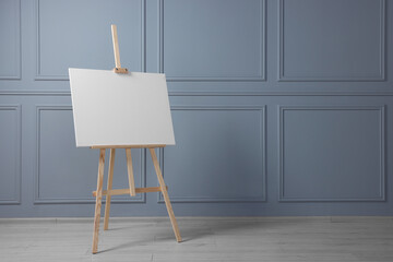 Wooden easel with blank canvas near grey wall indoors. Space for text