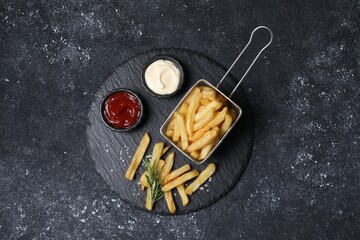 Tasty French fries, ketchup, rosemary and mayonnaise on black textured table, top view