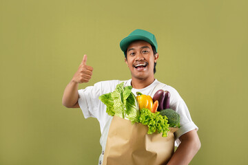 Photo of young asian man farm guy poses with organic veg box on green background in studio, smiles and shows thumb up