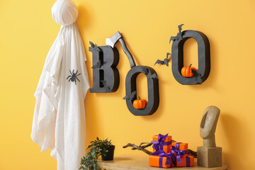 Table with gift boxes for Halloween, houseplant, ghost and text BOO hanging on orange wall in room, closeup