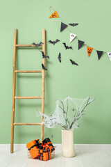 Vase with tree branches, gift boxes for Halloween, ladder and flags hanging on green wall in room