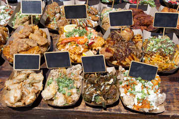 Close up on containers of fast food on brown table with blank label signs sticking into them. Fried Pickles, Cauliflower, grilled and with blue cheese and various flavors of chicken dishes. Fair food.