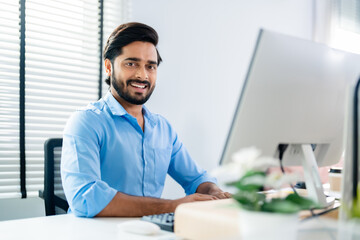 Indian professional business man, businessman working on computer, looks directly at camera and smiles friendly