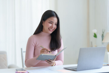 Smiling happy asian businesswoman sitting with laptop computer taking notes in office.