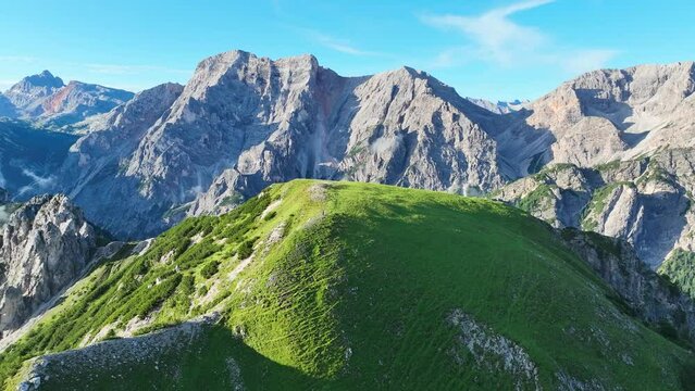 Scenic summer aerial view of a green hill in the foreground and the jagged Seekofel mountain peak in the background in Italy's Dolomite mountain range during the day.