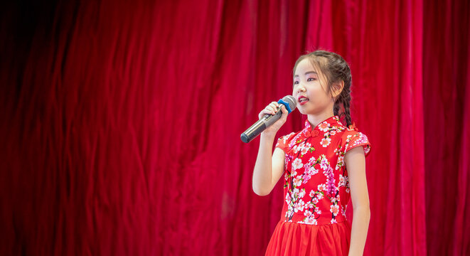 The Asian kid girl sing a song on stage at her school activity day, dress in Qipao style, red curtain background