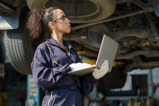 african american mechanic woman using laptop computer checking list to undercarriage of car in workshop at auto car repair service center with lift . car engineer female inspection vehicle details