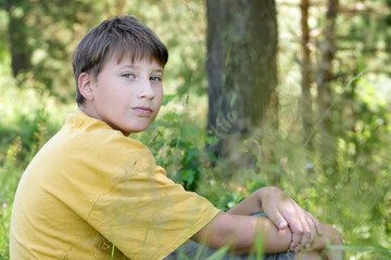 Teen boy sitting near a tree in the forest looking camera. Portrait of male 12-14 y.o. Copy space for a text
