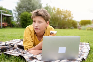Teen boy in yellow t-shirt laying the grass, eating ice cream, smiling and typing laptop in a park. 