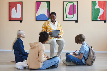 Group of teenagers sitting on floor in art gallery or museum and listening to lecture from black...