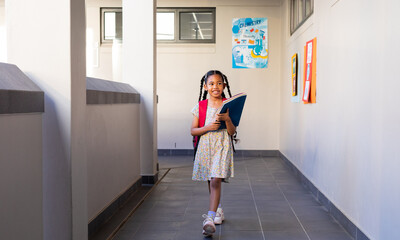 Happy biracial schoolgirl with books and school bag at elementary school corridor