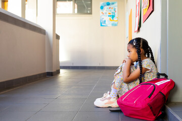 Sad biracial schoolgirl with school bag sitting at wall at elementary school corridor, copy space