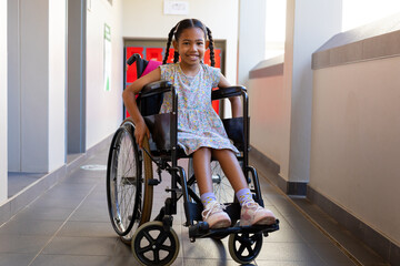 Fototapeta na wymiar Portrait of happy biracial schoolgirl sitting in wheelchair at school corridor at elementary school