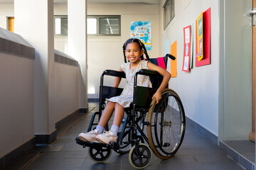 Fototapeta na wymiar Portrait of happy biracial schoolgirl sitting in wheelchair at elementary school corridor