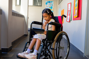 Focused biracial schoolgirl sitting in wheelchair and using tablet at elementary school corridor