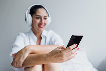 latin adult woman listening music with headphones and using mobile phone on bed at home in Mexico Latin America
