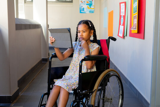 Focused biracial schoolgirl sitting in wheelchair and using tablet at elementary school corridor