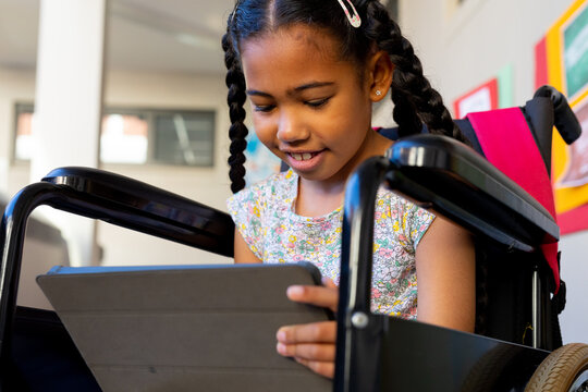 Happy biracial schoolgirl sitting in wheelchair and using tablet at elementary school corridor