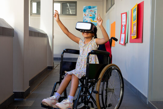 Happy biracial schoolgirl sitting in wheelchair and using vr headset at elementary school corridor