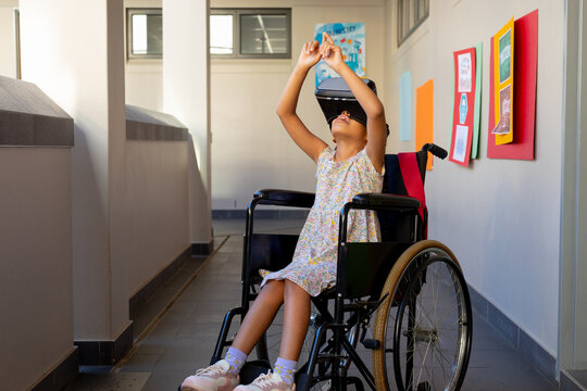 Happy biracial schoolgirl sitting in wheelchair and using vr headset at elementary school corridor