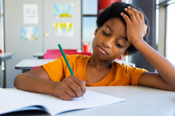Exhausted biracial schoolboy sitting at desk and writing in classroom